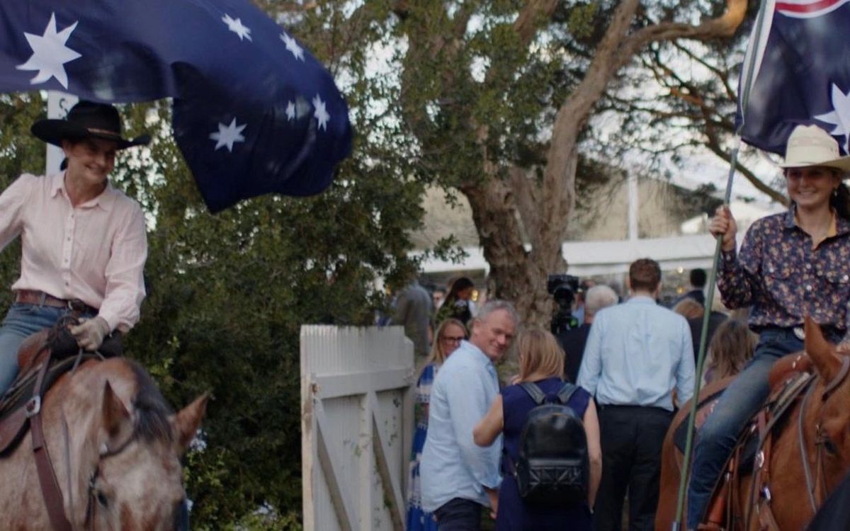 Two community members on horseback holding Australian flags, smiling among a gathered crowd outdoors