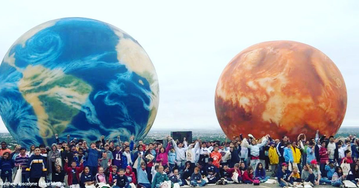 Community standing with large illuminated planetary spheres at dusk