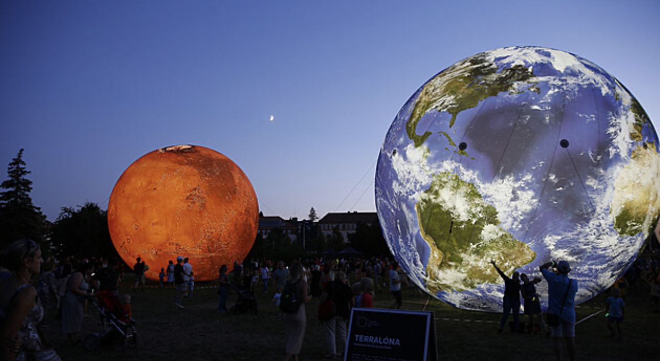 Community gathered at dusk around illuminated Earth and Mars spheres under a crescent moon